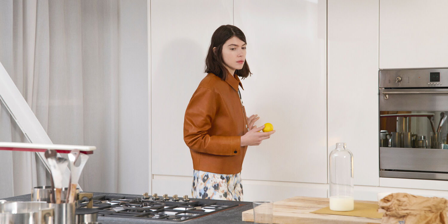 Model wearing a Marni brown leather jacket and printed skirt, holding a lemon in a modern kitchen.