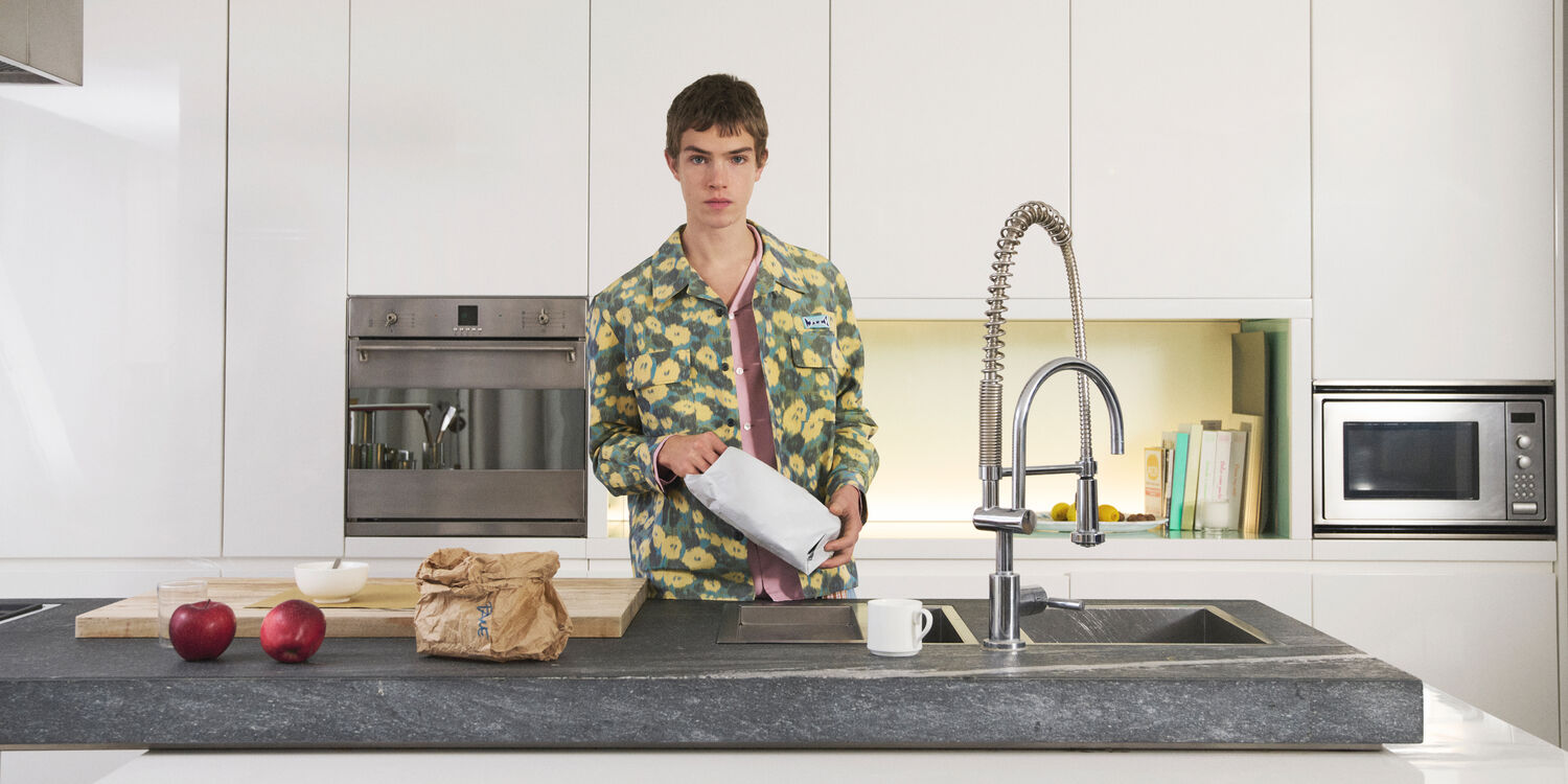 Model wearing a Marni floral jacket over a pink shirt in a modern kitchen, holding a paper bag.